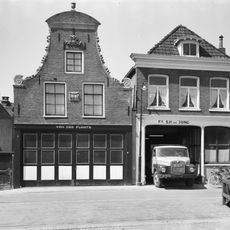 Pand onder zadeldak tegen klokgevel met gebeeldhouwde bekroning waarop siervaas