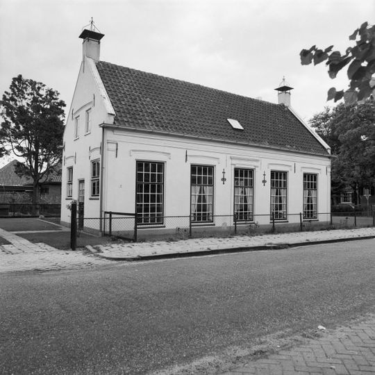 Gepleisterd dwarshuis onder zadeldak tussen puntgevels met hoekschoorstenen, waarop windkappen