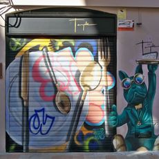 Mural que representa un plato y unos cubiertos junto a la figura de un perro con traje, pajarita y gafas multicolores que levanta el dedo pulgar de una de sus manos mientras con la otra sostiene una bandeja con un trozo de pastel.
