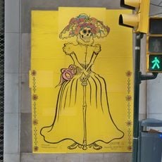 Póster compuesto por varias piezas de papel que forman un mosaico de color amarillo con el dibujo de un esqueleto vestido de mujer con traje y sombrero del siglo XIX apoyándose en un bastón hecho con huesos y una pequeña calavera.