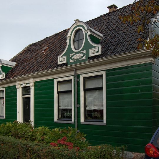 Houten huis met een ingang tussen Ionische pilasters en aan de straat tevens twee dakkapellen met ronde vensters onder gebogen kroonlijsten; zijmeanders en rozetten