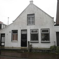 Woning met gepleisterde puntgevel onder zadeldak waarvan het linker schild dieper doorloopt dan het rechter