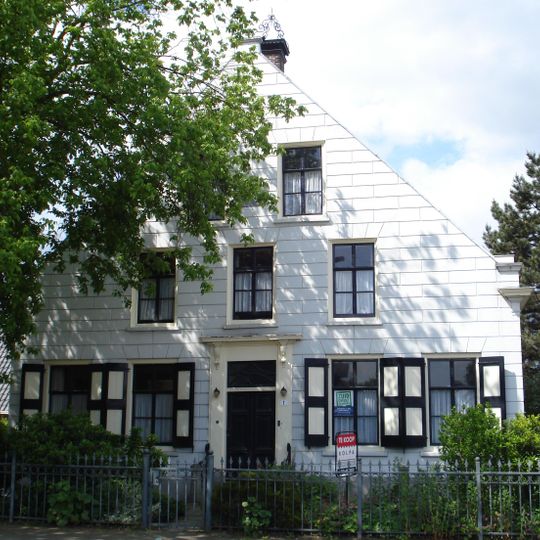 Boerderij met gepleisterd woonhuis onder zadeldak tussen puntgevels. Schoorstenen met smeedijzeren sierbekroningen. Vesters met luiken en zesruitsramen.