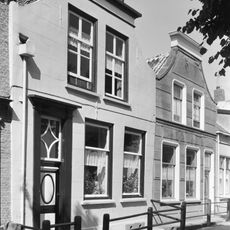 Huis met gepleisterde lijstgevel met ingezwenkte top. Schuifvensters. Voordeur met na-empire panelen en een omlijsting uit dezelfde periode