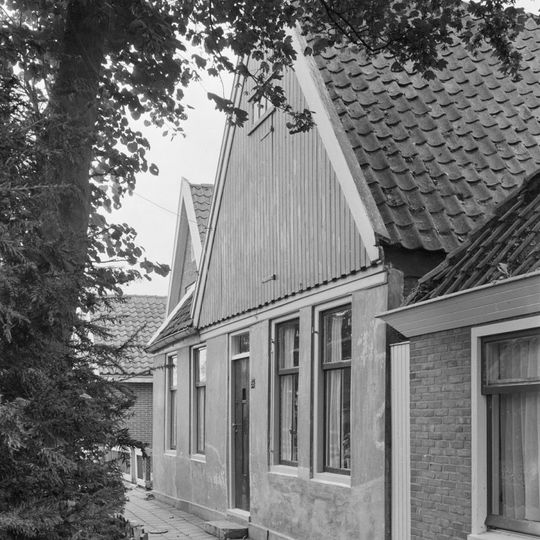 Pand onder zadeldak. Gepleisterde voorgevel waarboven een houten geveltopbeschieting. Aanbouw links