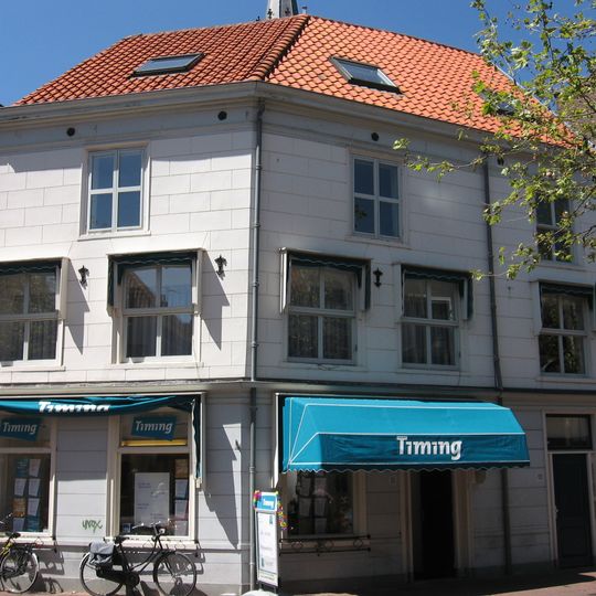 Hoekpand op geknikte plattegrond. Schilddak; gepleisterde lijstgevel met geprofileerde puibalk en oorspronkelijk deur